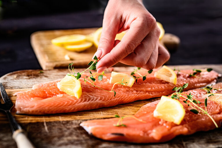 A person preparing baked salmon, placing lemon slices and fresh herbs on fillets.