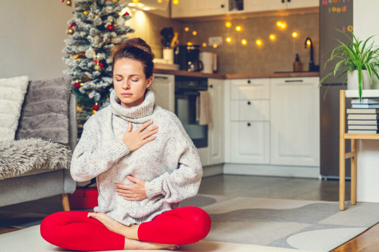 Woman practicing holiday mindfulness practices at home with hand on heart, near a Christmas tree